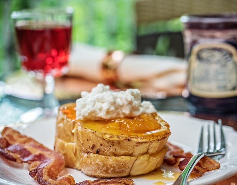 A plate of French toast topped with orange preserves and whipped cream, served with crispy bacon and accompanied by a glass of red beverage.