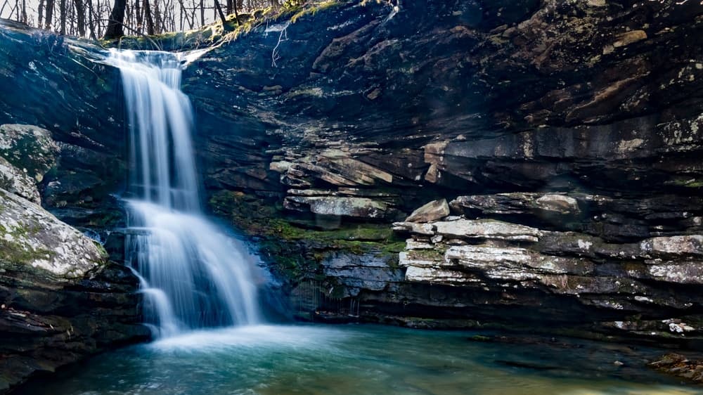 A serene waterfall cascades over rocky ledges into a tranquil pool surrounded by trees.