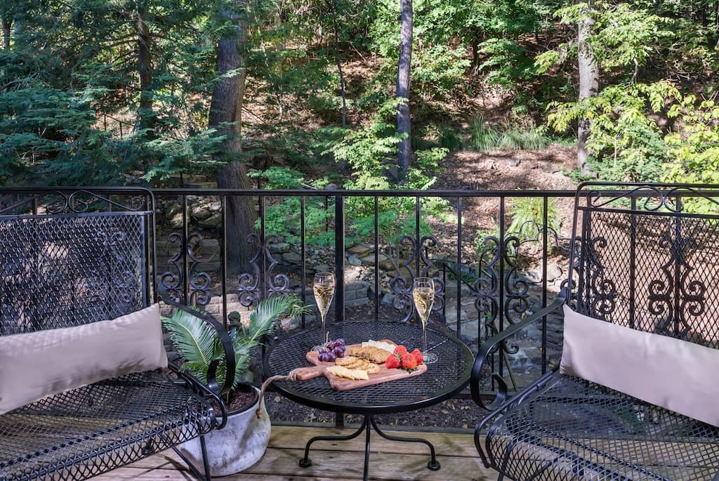 A cozy balcony with two chairs, a small table featuring fruit and snacks, and champagne glasses, surrounded by a lush, green forest.