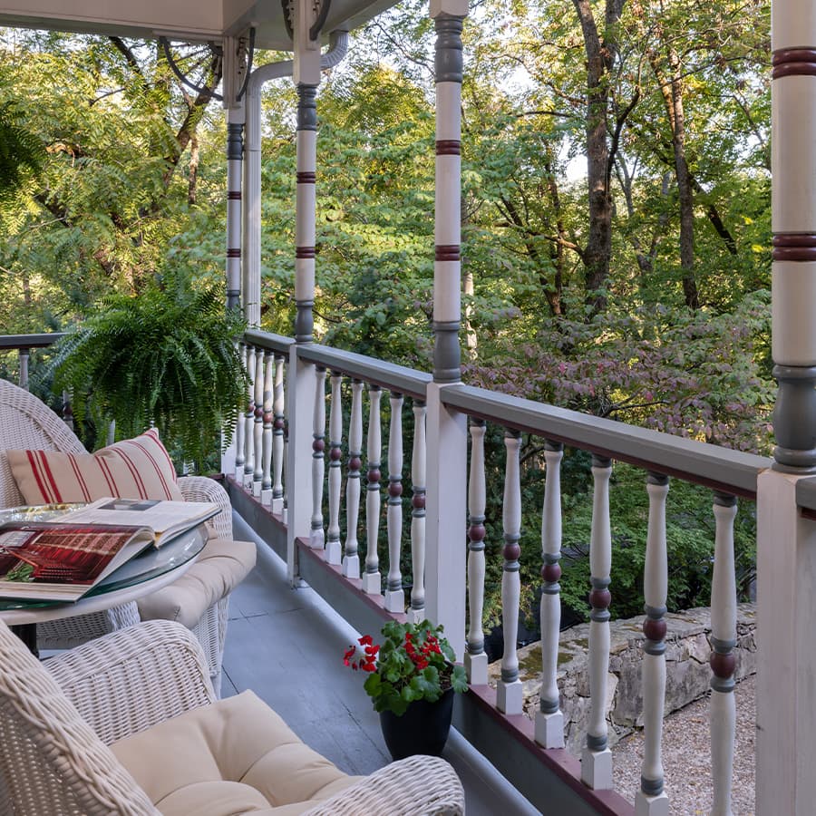 A spacious porch with white wicker seating, a small table, and a potted plant. The porch overlooks a lush green forest with tall trees and a stone wall.