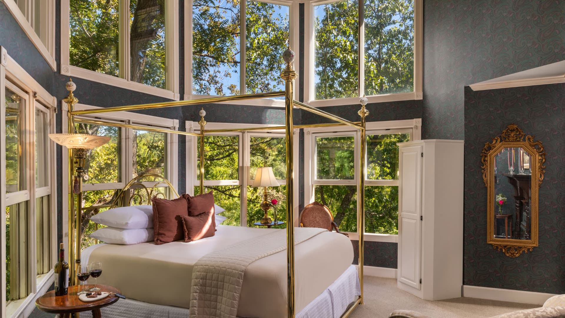 A spacious bedroom featuring a four-poster gold canopy bed with white linens and burgundy pillows. Large windows offer stunning views of the surrounding forest.