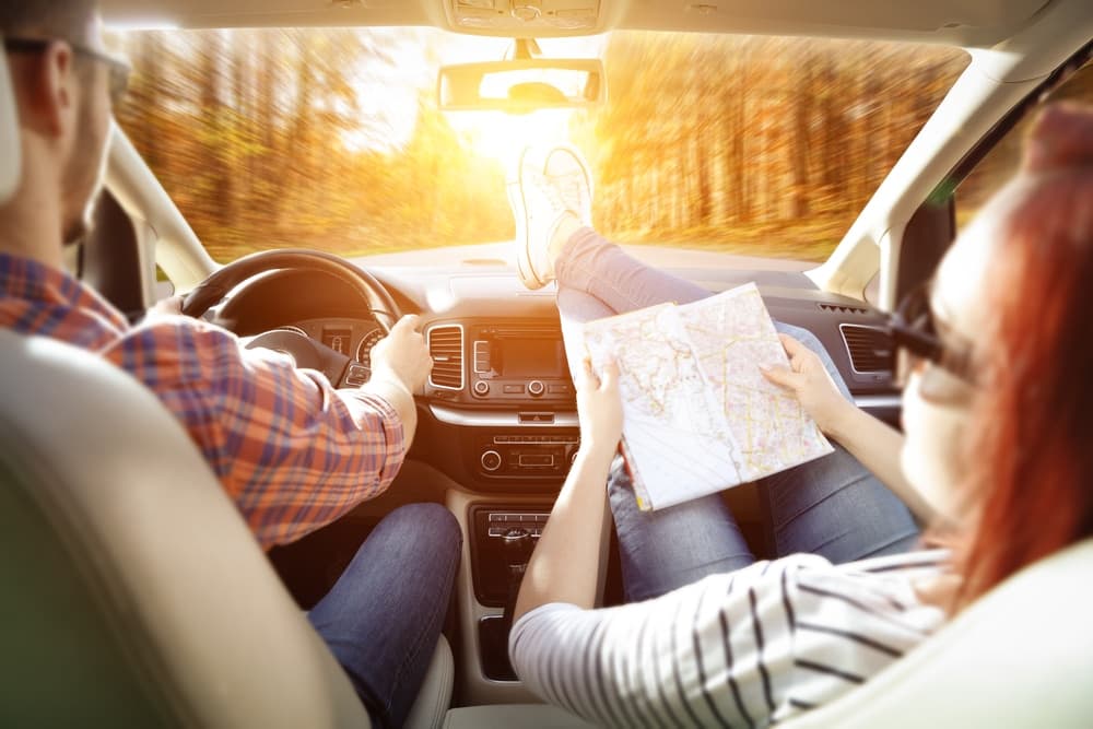 A couple sits in a car, with the driver steering and the passenger reading a map, enjoying a scenic drive.