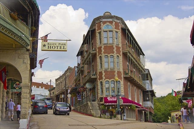 A sloped street leads to the historic Basin Park Hotel surrounded by shops and vibrant signage under a cloudy sky. A sloped street leads to the historic Basin Park Hotel surrounded by shops and vibrant signage under a cloudy sky.