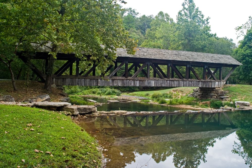 A wooden covered bridge spans a serene pond surrounded by lush greenery.