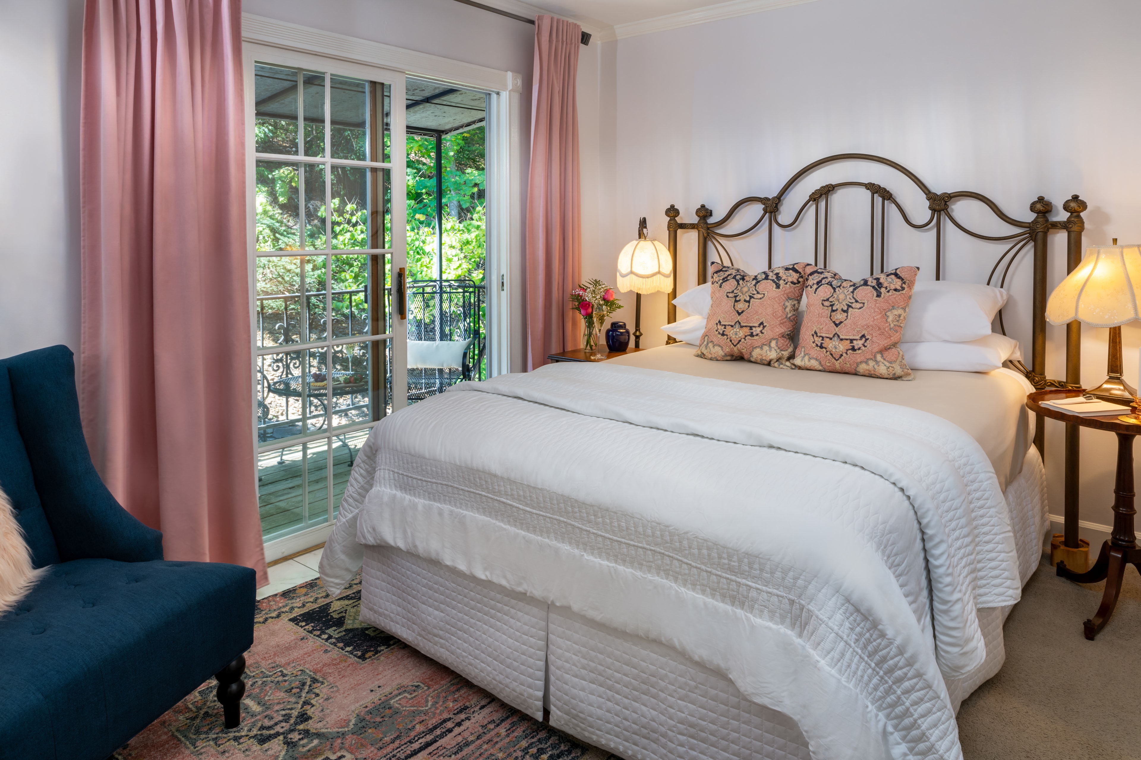 A cozy bedroom featuring a queen bed, pink curtains, a blue armchair, and a balcony door.