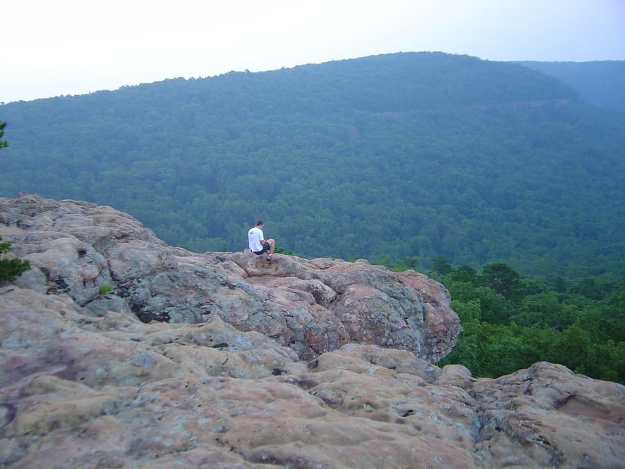 A person sits on a rocky ledge overlooking a dense green valley.