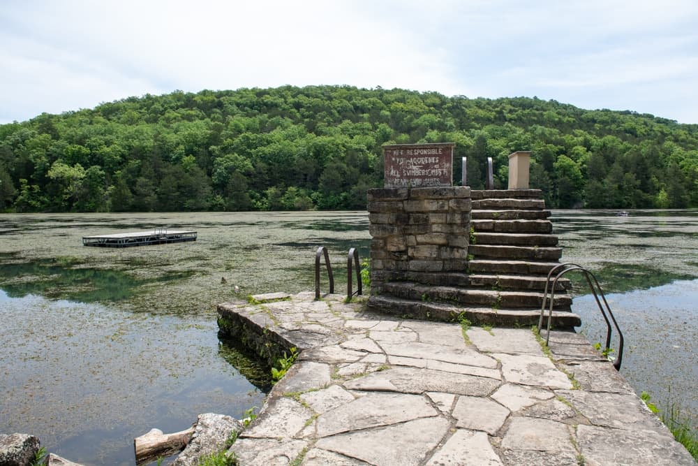 Stone steps lead down to a calm, green-tinted lake surrounded by lush hills.