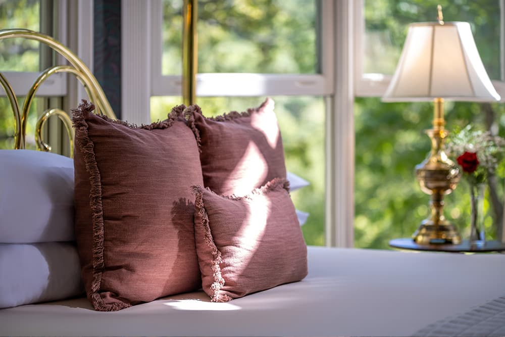 A bed with white sheets and a collection of soft, pink pillows. Sunlight streams through a window, casting a warm glow on the bed. A small table with a lamp and a vase of flowers sits nearby.