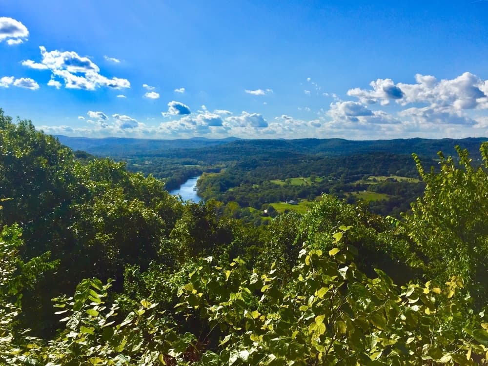 A panoramic view of a lush green valley with a winding river under a bright blue sky dotted with clouds.