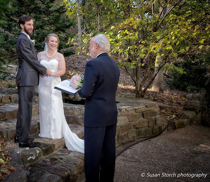 A bride and groom stand on stone steps smiling at each other while an officiant reads from a book in a wooded setting.