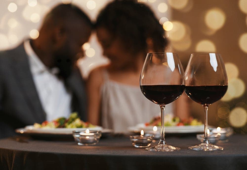A romantic dinner setting with two wine glasses in focus, beside a couple enjoying their meal. A romantic dinner setting with two wine glasses in focus, beside a couple enjoying their meal.