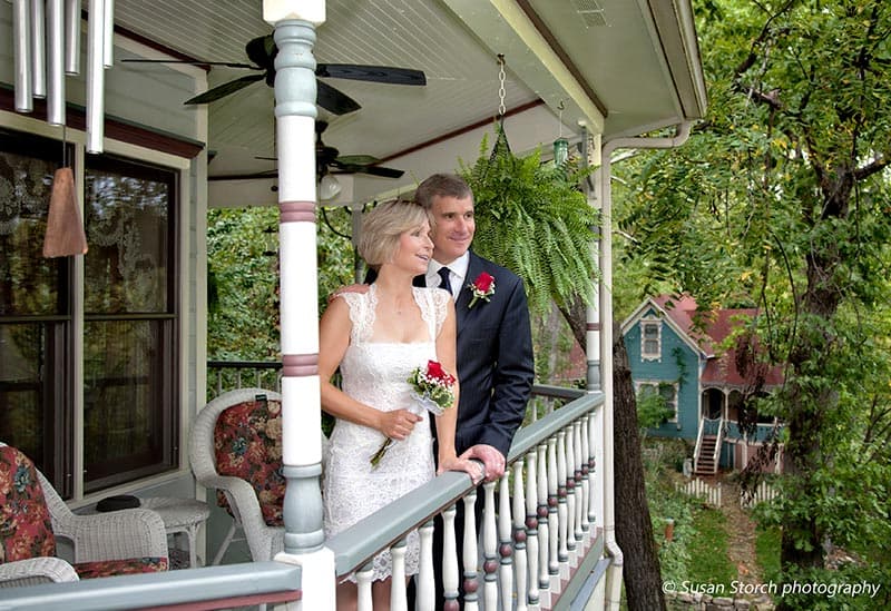 A happy couple stands on a porch, celebrating their wedding with greenery and colorful houses in the background.