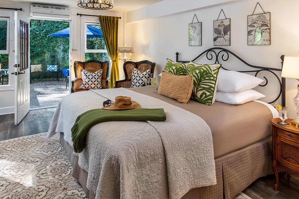 A luxurious bedroom with a king-size bed, featuring a wrought-iron headboard and soft bedding. The room is decorated with botanical-themed artwork and lush green plants. A pair of armchairs and a small table provide a comfortable seating area.