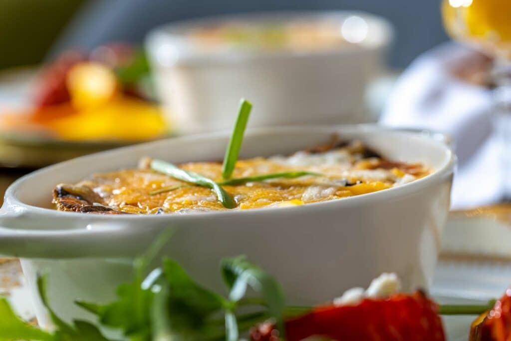A close-up of a baked dish in a white ramekin. The dish is topped with melted cheese and a sprig of chives.