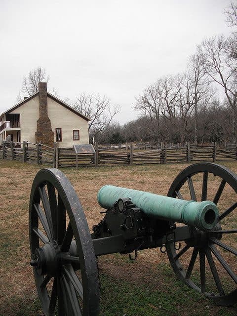 A green cannon stands in front of a historic building and a wooden fence under a cloudy sky.