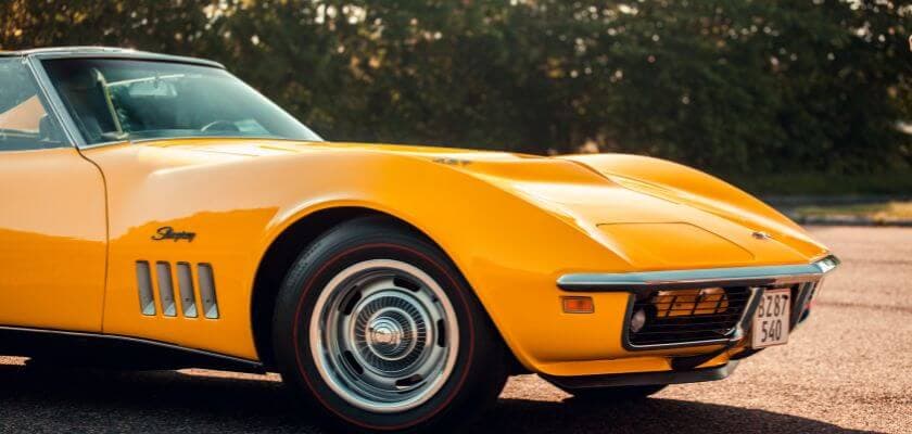 a yellow chevrolet stingray corvette with the soft top down parked on the pavement with a backdrop of lush green foliage of bushes and trees